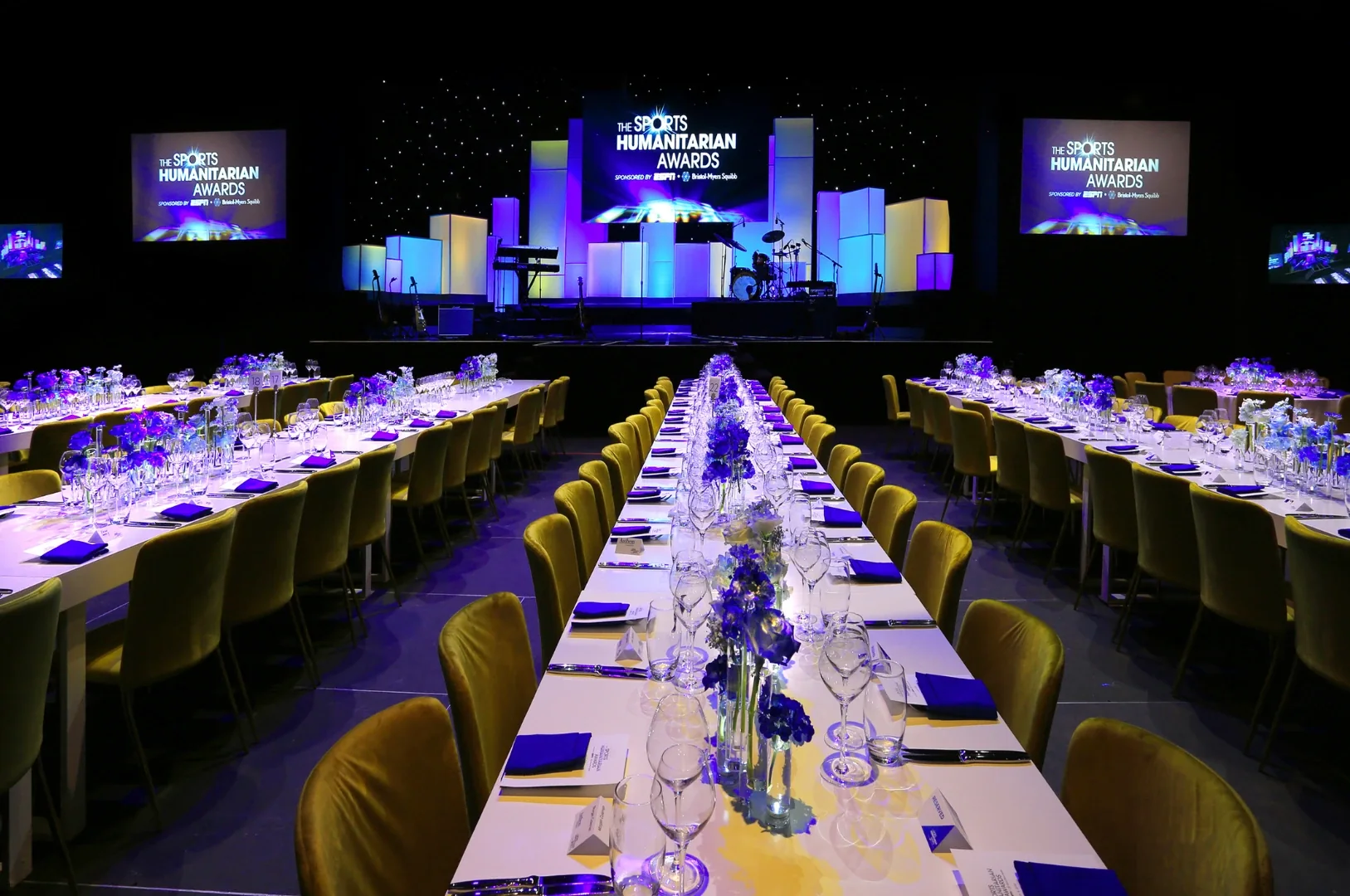 The Sports Humanitarian Awards dinner: long tables lit in blue and violet facing a glowing stage